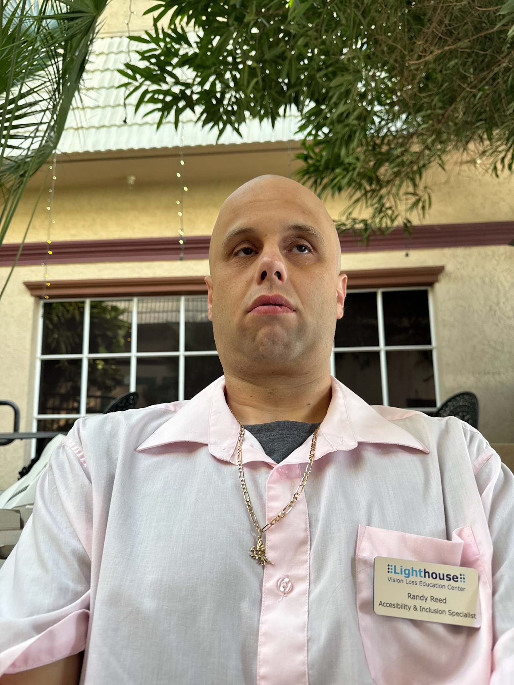 The picture shows a person from the chest up, standing outdoors. They are bald and looking slightly upwards towards the camera with a neutral expression. They are wearing a light pink button-up shirt with a grey t-shirt visible underneath. Around their neck, there is a gold chain with a small pendant. On their left chest, there is a name tag that reads "Lighthouse Vision Loss Education Center, Randy Reed, Accessibility & Inclusion Specialist." Behind them, there are green palm leaves at the top, a building with a yellow wall and red trim, and a window with black bars. There are also string lights hanging from the building's eaves.